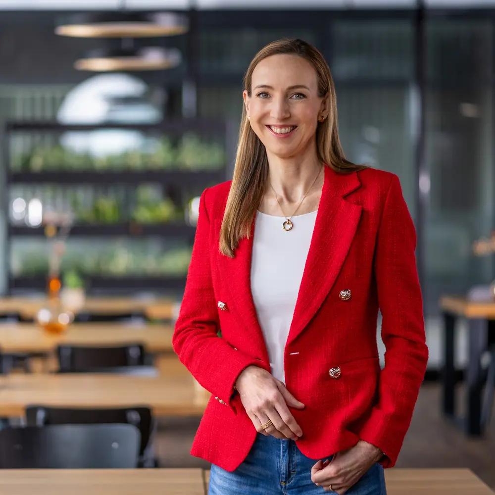 Η Δρ. Simone Bagel-Trah φοράει ένα έντονο κόκκινο σακάκι και λευκή μπλούζα, όρθια σε έναν μοντέρνο χώρο τύπου καφέ με ξύλινα τραπέζια και διακοσμητικά βάζα στο φόντο.